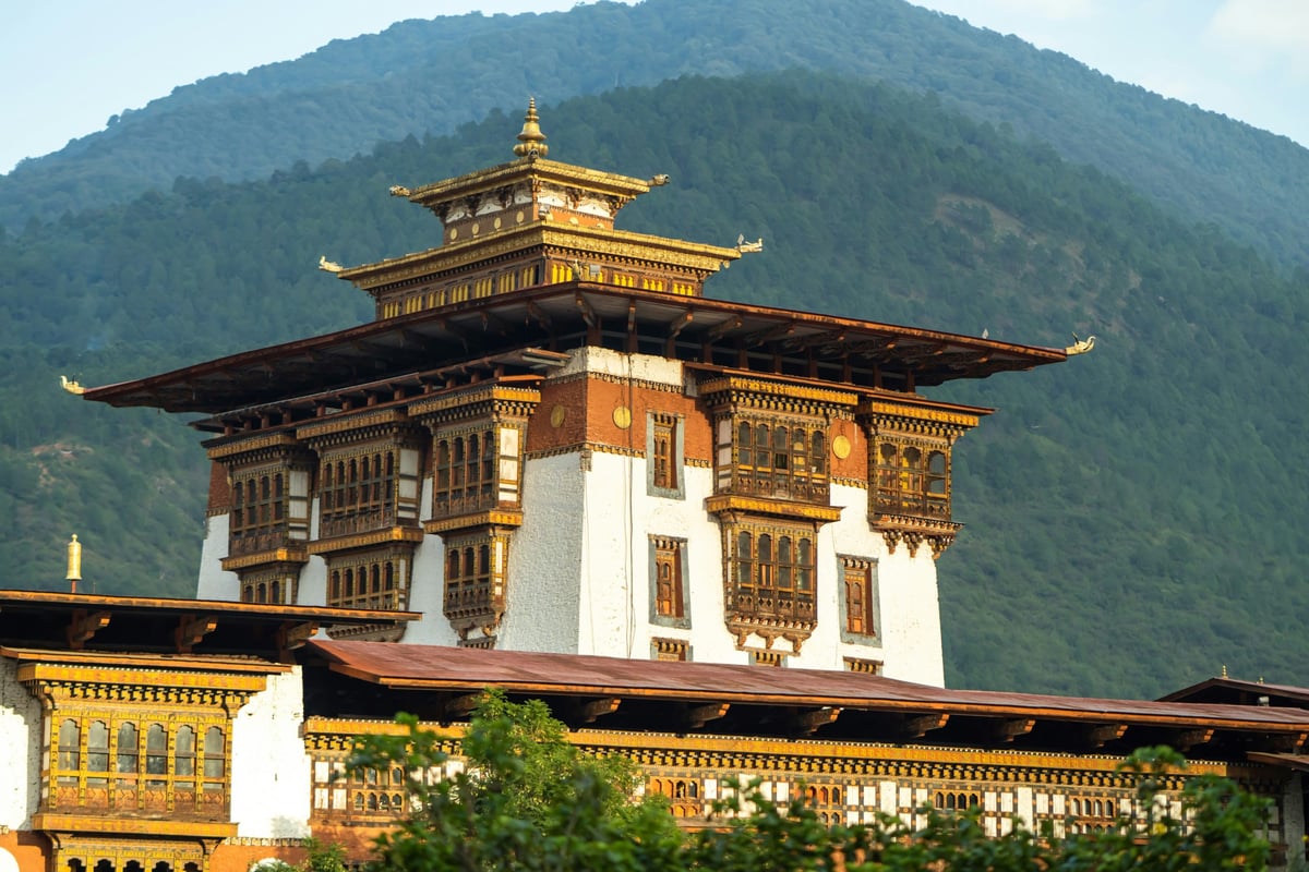 A Bhutanese monastery perched on a mountain, with a mix of white and red buildings, and a few trees in the foreground, with a mountainous background