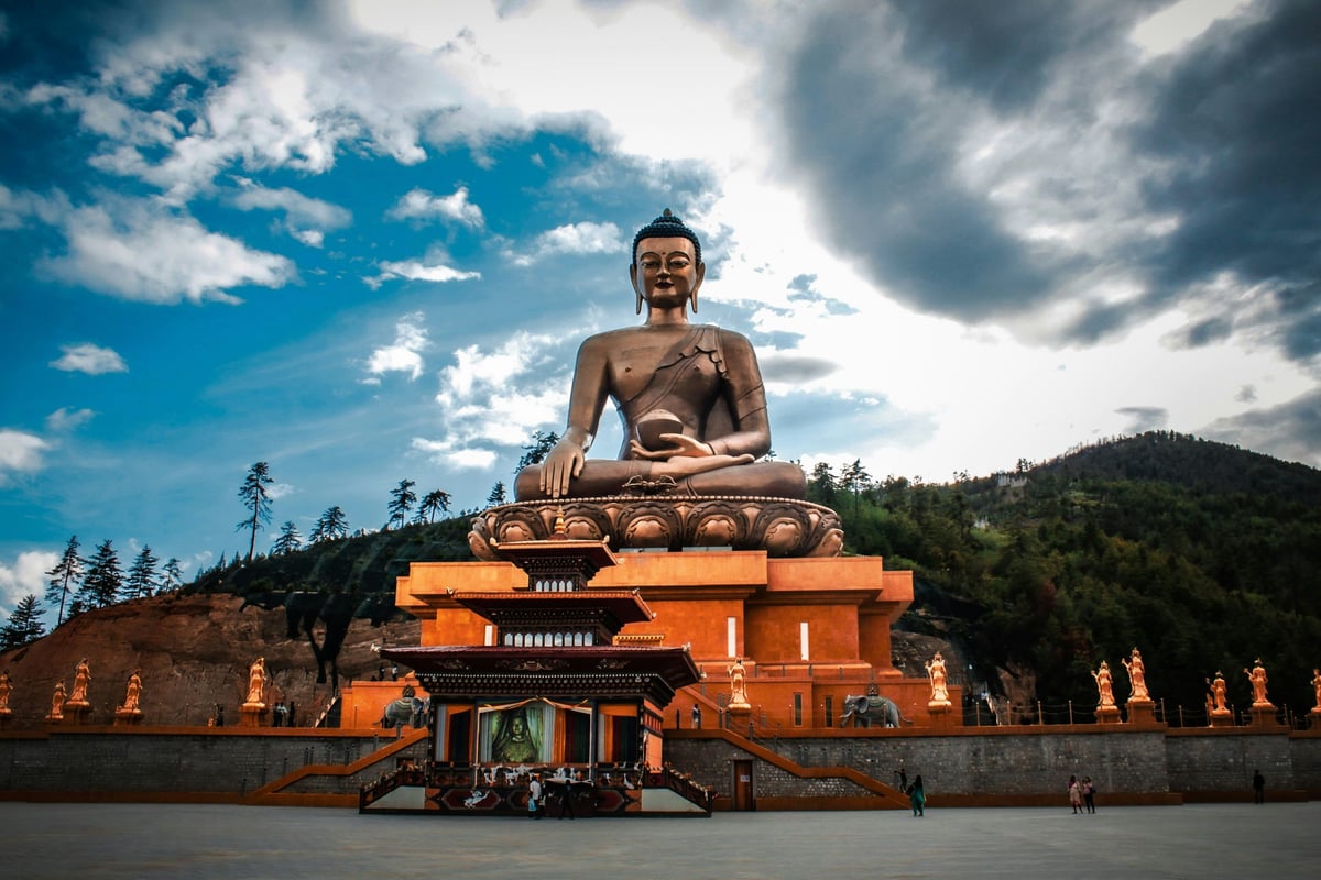 A large, brown, seated Buddha statue in front of a monastery in the mountains of Bhutan