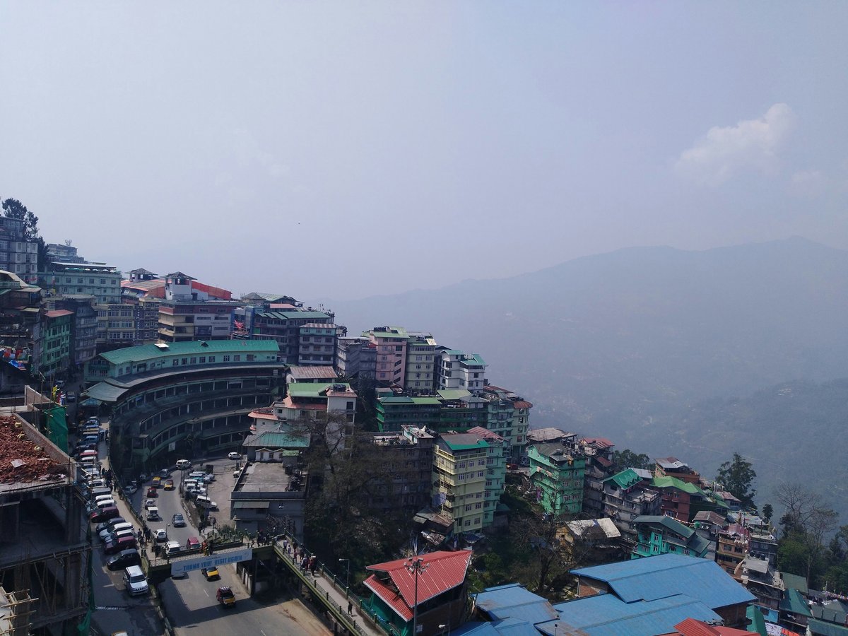 A small, hilly Gorkha in Nepal with a mix of old and new buildings, surrounded by mountains and trees, with a few people and vehicles on the streets