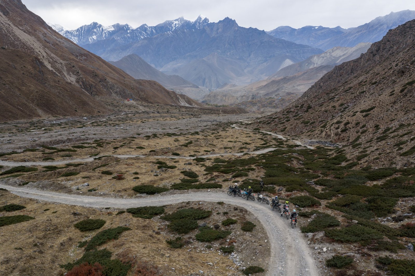 Group of motorcyclists riding along a winding dirt road in a high-altitude valley with snow-capped mountains in the background during our Nepal motorcycle tour.