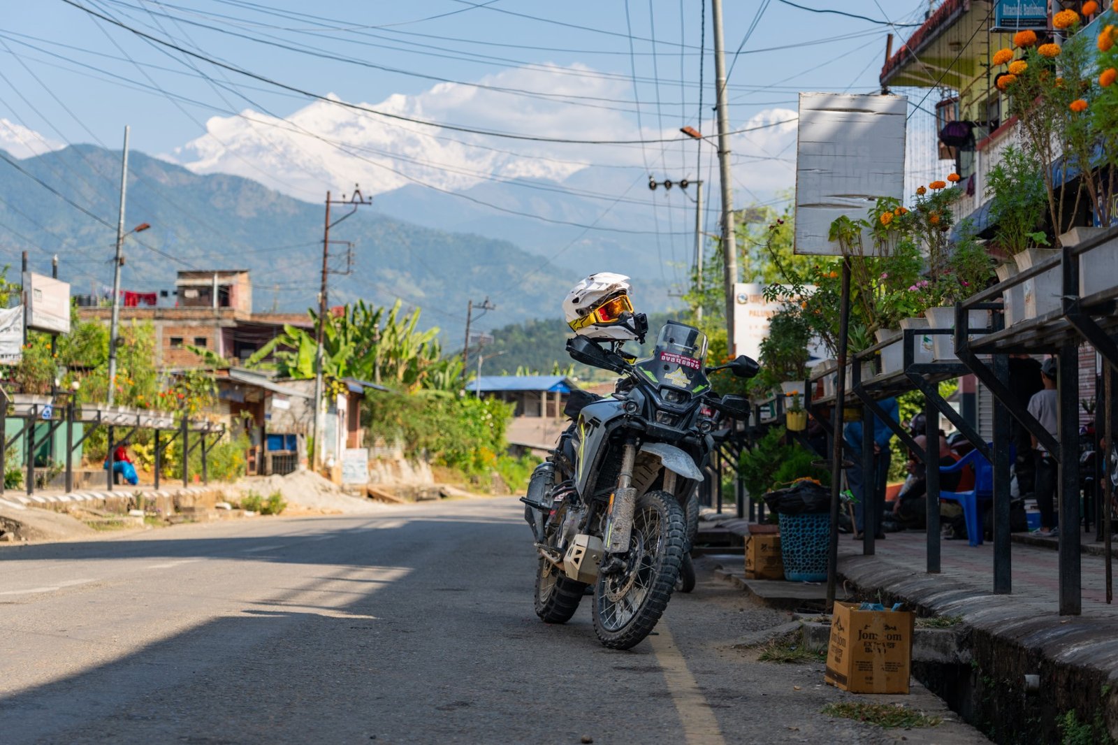 Adventure motorcycle parked on a village street with a marigold-adorned building and snow-capped mountains in the background during our Nepal motorcycle tour.