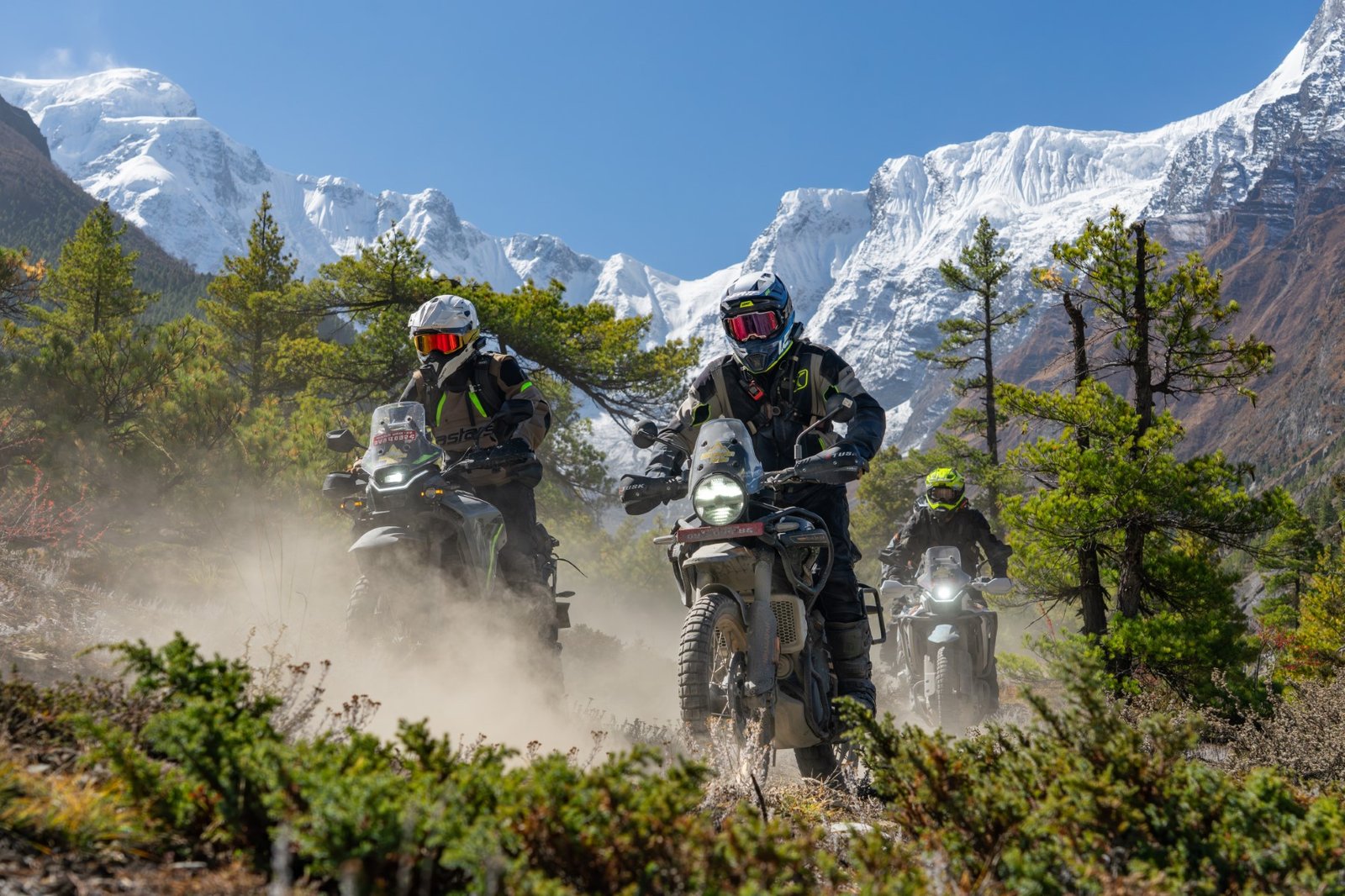 Three motorcyclists riding through a dusty forest trail with snow-capped mountains in the background during our Nepal motorcycle tour.
